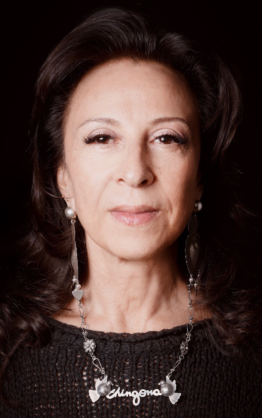 Image Description: Photo of a Mexican American woman facing the camera. She has shoulder-length brown hair and wears long silver earrings and a silver chain necklace that says “Chingona” framed by two birds. The necklace rests on the front of her black sweater. Photo Credit: Kevin Abosch