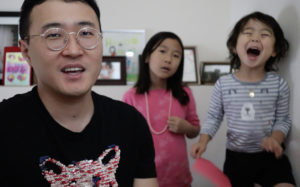 Image Description: Photo of a Korean American man with short black hair and gold-rimmed glasses, looking into the camera. Behind him are two children. One of the children has long black hair and a pink shirt, and is looking inquisitively at the camera. The other child is wearing a striped gray and white shirt, and has their mouth open and eyes closed in excitement. Photo Credit: Matthew Salesses