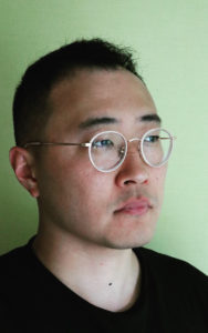 Photo of a Korean American man with short black hair, wearing gold rimmed glasses and a black shirt, standing against a green wall. He is looking past the camera. Photo Credit: Matthew Salesses