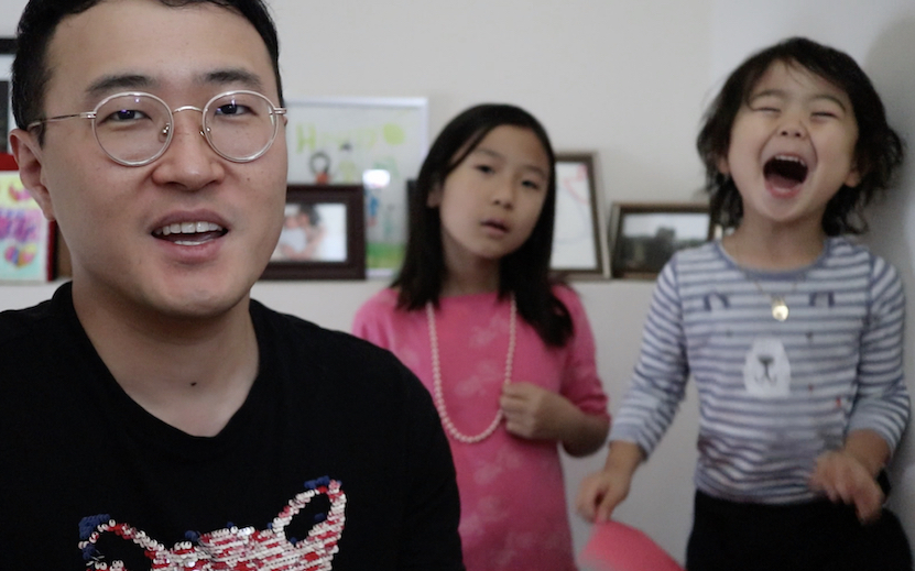 [Image Description: Photo of a Korean American man with short black hair and gold-rimmed glasses, looking into the camera. Behind him are two children. One of the children has long black hair and a pink shirt, and is looking inquisitively at the camera. The other child is wearing a striped gray and white shirt, and has their mouth open and eyes closed in excitement.] Photo Courtesy of Matthew Salesses
