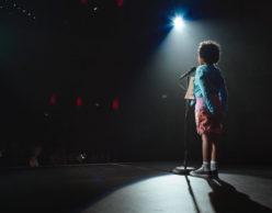 [Image Description: Photo of a child standing on a stage with a microphone in front of them and their hands clasped behind their back. They are standing beneath a spotlight with their back to the camera as they face the front of the stage. They have short, dark curly hair, and they are wearing a pale blue shirt, red shorts, and sneakers. The rest of the image is dark.] Photo Credit: June Street Productions