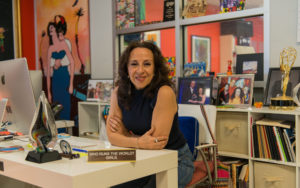 Image Description: Photo of a Mexican American woman seated at her work desk and leaning forward while smiling at the camera. She has layered brown hair that rests below her shoulders and wears jeans, a sleeveless navy blue shirt, and silver earrings. The shelf behind her holds framed photographs, artwork, and books. On her desk is a nameplate that reads “Who runs the world? Girls.” Photo Credit: Columbia Spectator, Michael Cao