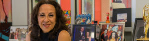 Image Description: Photo of a Mexican American woman smiling at the camera. She has layered brown hair that rests below her shoulders and wears a sleeveless navy blue shirt, and silver earrings. Behind her are numerous framed photographs, artwork, and a gold award statue. Photo Credit: Columbia Spectator, Michael Cao