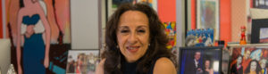 [Image Description: Photo of a Mexican American woman smiling at the camera. She has layered brown hair that rests below her shoulders and wears a sleeveless navy blue shirt, and silver earrings. Behind her are colorful framed photographs and artwork.] Photo Credit: Columbia Spectator, Michael Cao