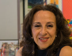 [Image Description: Photo of a Mexican American woman smiling at the camera. She has layered brown hair that rests below her shoulders and wears a sleeveless navy blue shirt, and silver earrings. Behind her are colorful framed photographs and artwork.] Photo Credit: Columbia Spectator, Michael Cao
