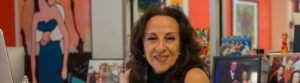 [Image Description: Photo of a Mexican American woman smiling at the camera. She has layered brown hair that rests below her shoulders and wears a sleeveless navy blue shirt, and silver earrings. Behind her are colorful framed photographs and artwork.] Photo Credit: Columbia Spectator, Michael Cao