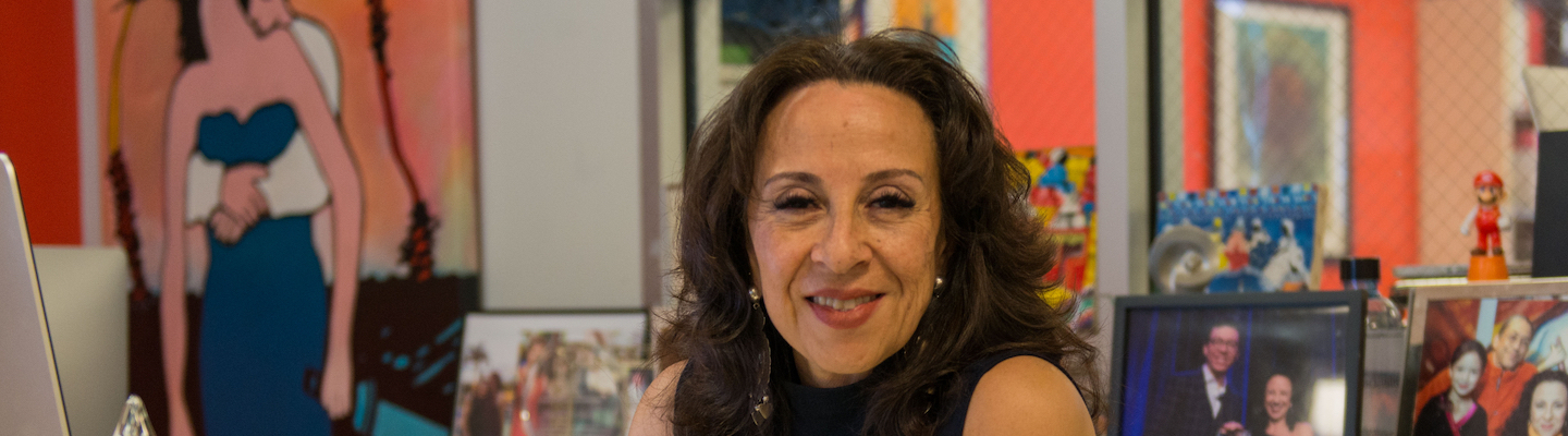 [Image Description: Photo of a Mexican American woman smiling at the camera. She has layered brown hair that rests below her shoulders and wears a sleeveless navy blue shirt, and silver earrings. Behind her are colorful framed photographs and artwork.] Photo Credit: Columbia Spectator, Michael Cao