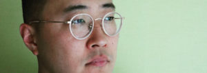 [Image Description: Photo of a Korean American man with short black hair, wearing gold rimmed glasses and a black shirt, standing against a green wall. He is looking past the camera.] Photo Courtesy of Matthew Salesses