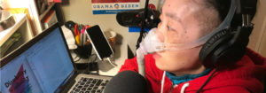 [Image Description: Photo of an Asian American woman at a desk. She is looking at her laptop computer and there is a microphone in front of her. She has headphones over her ears and is wearing a mask over her nose attached to a gray tube.] Photo Courtesy of Alice Wong
