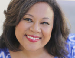 [Image Description: Photo of a Black woman smiling into the camera. She has shoulder-length brown hair with gold highlights, and is wearing red lipstick and a purple shirt with a zig-zag pattern. The sunlight is shining on the side of her face.] Photo Credit: Andrea Scher