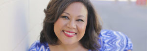 [Image Description: Photo of a Black woman smiling into the camera. She has shoulder-length brown hair with gold highlights, and is wearing red lipstick and a purple shirt with a zig-zag pattern. The sunlight is shining on the side of her face.] Photo Credit: Andrea Scher