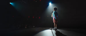 [Image Description: Photo of a child standing on a stage with a microphone in front of them and their hands clasped behind their back. They are standing beneath a spotlight with their back to the camera as they face the front of the stage. They have short, dark curly hair, and they are wearing a pale blue shirt, red shorts, and sneakers. The rest of the image is dark.] Photo Credit: June Street Productions