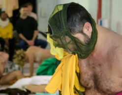 [Image Description: Photo of a Latinx man in the middle of a performance. He is shirtless and has a green pair of mesh underwear over his face as he looks down towards the ground. Bright pink and yellow pieces of fabric are wrapped around his arms. In the background are two other performers, one sitting on the ground and one on all fours. Behind them are seated audience members observing the performance. There is fabric and other items strewn across the performance space floor.] Photo Credit: Ian Douglas